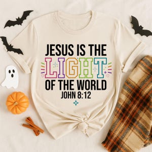 May include: A cream-colored short-sleeve t-shirt featuring the black text "JESUS IS THE LIGHT OF THE WORLD JOHN 8:12" with the word "LIGHT" printed in large, colorful, outlined block letters, accented by small star, heart, and plus symbols.