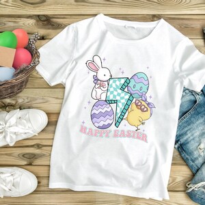 Puede incluir: Camiseta blanca con un gr&aacute;fico de un conejito, un pollito y huevos de Pascua. El gr&aacute;fico incluye un rayo y el texto "Feliz Pascua".