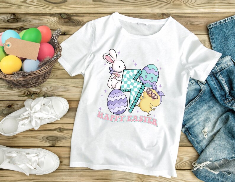 Puede incluir: Camiseta blanca con un gr&aacute;fico de un conejito, un pollito y huevos de Pascua. El gr&aacute;fico incluye un rayo y el texto "Feliz Pascua".