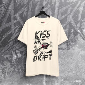 May include: A cream colored t-shirt with a black and white graphic print of a woman's lips with lipstick and a lipstick tube. The text "Kiss My Drift" is printed on the shirt. The text "Kiss My Drift" is printed in black and the text "Drift" is printed in a larger font size than the text "Kiss My".