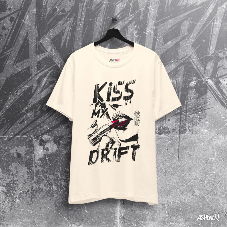 May include: A cream colored t-shirt with a black and white graphic print of a woman's lips with lipstick and a lipstick tube. The text "Kiss My Drift" is printed on the shirt. The text "Kiss My Drift" is printed in black and the text "Drift" is printed in a larger font size than the text "Kiss My".
