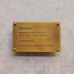 Puede incluir: Placa rectangular de latón con la palabra "Versus" y detalles del producto. La placa está asegurada con cuatro pequeños tornillos dorados. El texto incluye "Materials: Patina Brass, Aged Steel, Urban Bronze" y "Socket Based."
