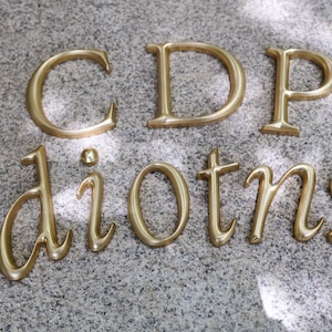 以下が含まれることがあります： 「CDP diotn」と綴られた金色の金属文字と数字のクローズアップ。文字はセリフ体で、まだらの灰色の表面に配置されています。照明は金属の表面にハイライトと影を作り出しています。