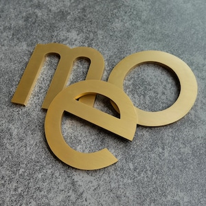 May include: Three gold-coloured metal letters, spelling "me", are arranged on a grey, textured surface. The letter "m" is positioned at the top left, the "e" in the bottom centre, and the "o" is overlapping the "e".