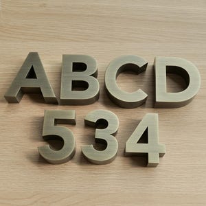 Puede incluir: Letras y números de metal color bronce dispuestos sobre una superficie de madera clara. Se muestran las letras A, B, C y D, junto con los números 5, 3 y 4. Los objetos tienen una estética vintage e industrial.