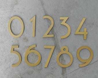 Könnte beinhalten: Ein Satz von zehn messingfarbenen Hausnummern aus Metall, von Null bis Neun, auf einer grauen Oberfläche angeordnet. Jede Zahl ist in einer einfachen, modernen Schriftart gehalten. Die Zahlen sind wahrscheinlich für ein Haus oder einen Briefkasten.
