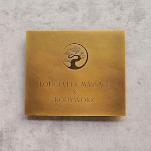 Puede incluir: Un letrero cuadrado de color dorado con las palabras "LONGEVITY MASSAGE & BODYWORK" en una fuente serif. Encima del texto hay un logotipo circular con un árbol estilizado. El letrero tiene una superficie texturizada.