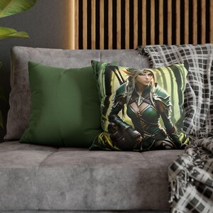 May include: Two cushions on a grey sofa. One is plain green, the other has a fantasy illustration of a female elf in green armour, set against a forest background. A plaid blanket is draped over the sofa, adding a touch of comfort.