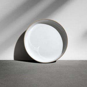 Könnte beinhalten: Ein weißer Keramikteller mit leicht erhöhtem Rand, vor einem neutralgrauen Hintergrund. Der Teller ist angewinkelt, was sein klares, minimalistisches Design hervorhebt. Schatten erzeugen ein Muster an der Wand hinter dem Teller.