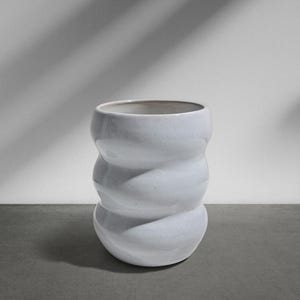 Könnte beinhalten: Eine weiße Keramikvase mit einem einzigartigen, gestapelten, abgerundeten Design. Die Vase hat eine glatte, glänzende Oberfläche und eine breite Öffnung. Die Vase ist etwa 25 cm hoch und 20 cm breit. Der Hintergrund ist eine schlichte weiße Wand mit Schatten.