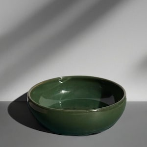 Könnte beinhalten: Eine glänzende, tiefgrüne Keramikschale. Die Schale hat eine abgerundete Form mit einem leicht ausgestellten Rand. Das Innere reflektiert das Licht, und die Außenseite hat eine glatte, gleichmäßige Oberfläche. Die Schale steht auf einer grauen Oberfläche mit Schatten.