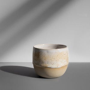 Könnte beinhalten: Eine handgefertigte Keramiktasse mit abgerundeter Form. Die Tasse hat ein zweifarbiges Design mit einem hellbeigen Boden und einem gesprenkelten weißen und beigen oberen Bereich. Die Tasse ist für Getränke konzipiert.