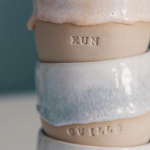 May include: Stacked ceramic mugs in shades of white, pink, and blue. The mugs have a textured, organic appearance with the word "KUN" and "GUILL" stamped on the sides. The mugs are handmade and have a rustic aesthetic.