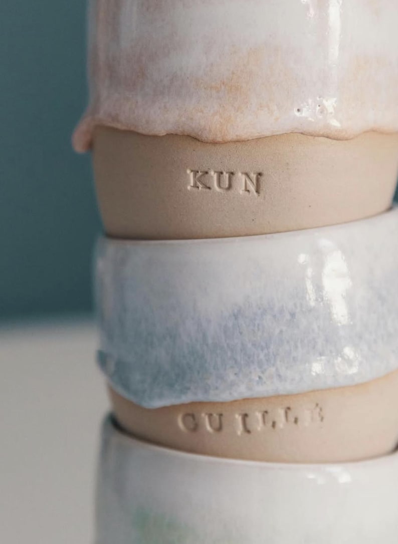 May include: Stacked ceramic mugs in shades of white, pink, and blue. The mugs have a textured, organic appearance with the word "KUN" and "GUILL" stamped on the sides. The mugs are handmade and have a rustic aesthetic.