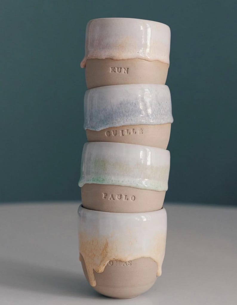 May include: A stack of five handmade ceramic cups. Each cup has a unique glaze color, including pink, blue, green, and tan, with a dripped effect. The names "KUN", "GUILLA", "PAULO", and "OMAS" are inscribed on the cups.
