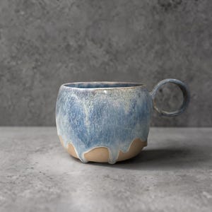 Puede incluir: Una taza de cerámica con cuerpo redondeado y asa circular. La parte superior presenta un esmalte azul y blanco moteado que gotea sobre la sección inferior, la cual es de arcilla color canela sin esmaltar con un acabado mate y suave.
