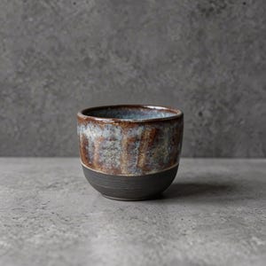 Könnte beinhalten: Eine handgefertigte Keramiktasse mit dunkelgrauem Boden und einer strukturierten, glasierten oberen Hälfte. Die Glasur weist eine Mischung aus Braun-, Blau- und Weißtönen auf und erzeugt ein einzigartiges, handwerkliches Aussehen. Der Rand der Tasse ist heller.