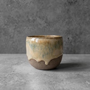 May include: A ceramic cup with a rounded shape, featuring a smooth, glossy glaze in shades of cream, beige, and soft blue that drips down over a textured, dark brown, unglazed base. The cup has a wide, open top and a sturdy, curved body.
