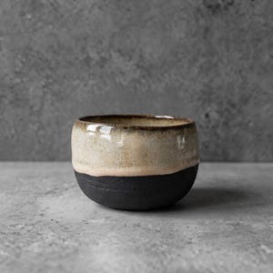 Puede incluir: Un cuenco de cerámica pequeño y redondo con un diseño de dos tonos. La parte superior tiene un esmalte suave y brillante en color crema y beige con sutiles motas marrones, mientras que la parte inferior tiene un acabado negro mate y texturizado.