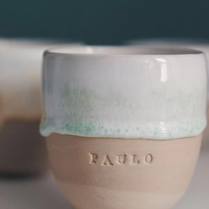 May include: A ceramic mug with a white top and a light brown base. The top has a subtle green tint where the white glaze meets the base. The name "PAULO" is embossed on the base. The mug is designed with a minimalist aesthetic.
