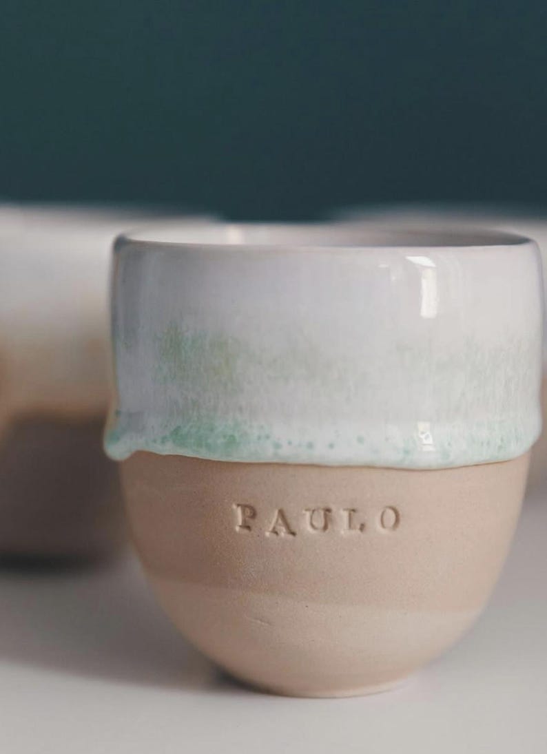 May include: A ceramic mug with a white top and a light brown base. The top has a subtle green tint where the white glaze meets the base. The name "PAULO" is embossed on the base. The mug is designed with a minimalist aesthetic.
