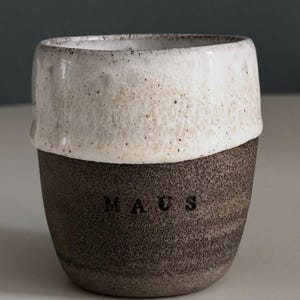 May include: A handcrafted ceramic mug with a two-tone design. The top half is white with a speckled texture, and the bottom half is dark brown with the word "MAUS" embossed. A unique, rustic style.