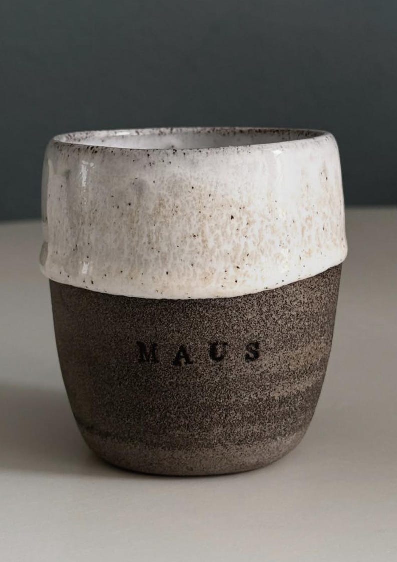 May include: A handcrafted ceramic mug with a two-tone design. The top half is white with a speckled texture, and the bottom half is dark brown with the word "MAUS" embossed. A unique, rustic style.