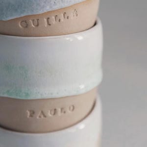 May include: A stack of handmade ceramic bowls. The top bowl has a light blue glaze, the second bowl has the name "GUILLA" etched into the clay. The third bowl is white with a hint of green, and the fourth bowl has the name "PAULO" etched into the clay.