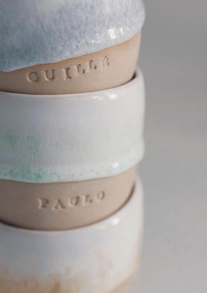 May include: A stack of handmade ceramic bowls. The top bowl has a light blue glaze, the second bowl has the name "GUILLA" etched into the clay. The third bowl is white with a hint of green, and the fourth bowl has the name "PAULO" etched into the clay.