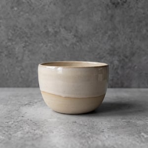 Könnte beinhalten: Eine Keramikschale mit abgerundeter Form, die eine glatte, glänzend cremeweiße Glasur im oberen Bereich und ein mattes, naturbeiges Finish im unteren Bereich aufweist, was ein zweifarbiges Erscheinungsbild mit einer sichtbaren Trennlinie erzeugt.