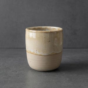 Könnte beinhalten: Eine handgefertigte Keramiktasse mit beiger Glasur. Die Tasse hat eine abgerundete Form mit leicht strukturierter Oberfläche und einem helleren Band in der Mitte. Der Innenraum ist sichtbar und scheint die gleiche Farbe wie die Außenseite zu haben.