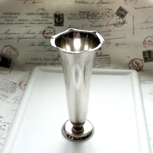Könnte beinhalten: Eine silberfarbene Vase mit einem ausgestellten, gewellten Rand und einem schlanken, sich verjüngenden Körper. Die Vase steht auf einem kleinen, kreisförmigen Sockel. Der Hintergrund zeigt Vintage-Papier mit verblasstem Text und Stempeln. Die Vase ist etwa 20 cm hoch.
