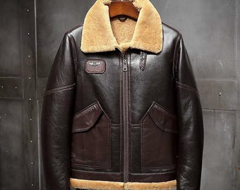 Puede incluir: Una chaqueta de vuelo de cuero marrón oscuro con cuello, puños y dobladillo de borreguillo beige grueso. La prenda cuenta con un cierre de cremallera central, dos grandes bolsillos delanteros con solapa y un parche rectangular en el pecho.
