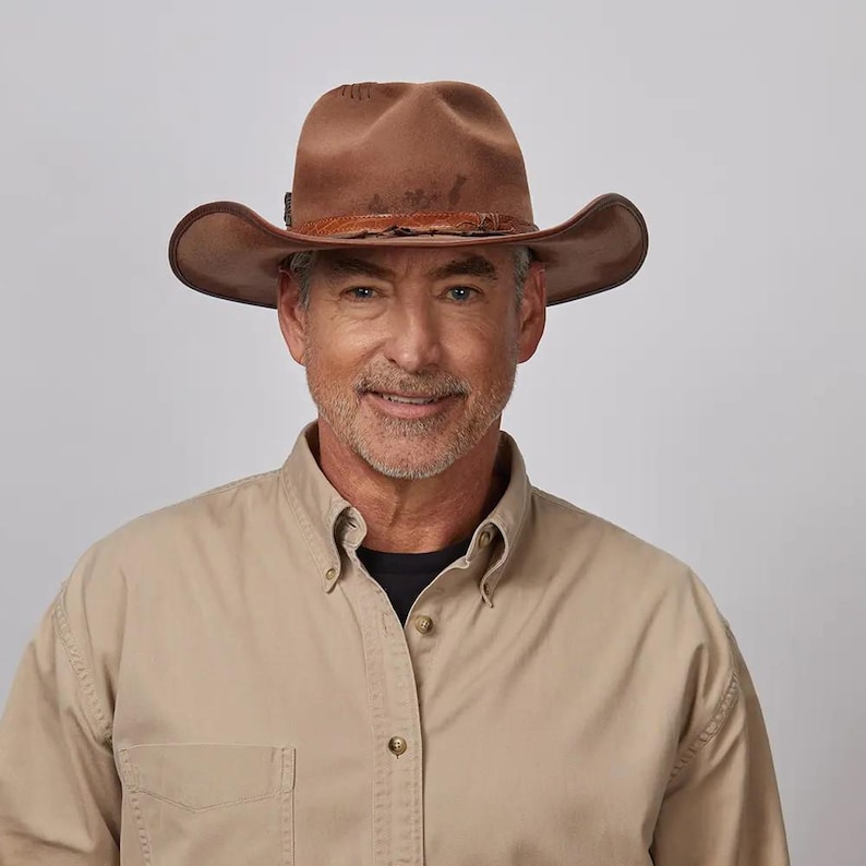 May include: A brown felt cowboy hat featuring a textured leather hatband with a distressed finish. The hat has a pinched crown and a wide, curved brim. The material appears smooth with subtle variations in color across the surface of the crown.