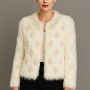 May include: Ivory faux fur jacket with pearl and embroidered embellishments. The jacket features a round collar and long sleeves. The design includes pearl accents and embroidered diamond patterns. The model is wearing black pants.