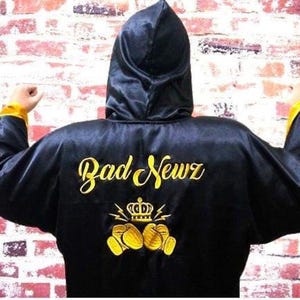 May include: Black satin boxing robe with a hood and yellow trim. The back of the robe features the embroidered text "Bad Newz" in gold script, along with a crown and boxing glove design.