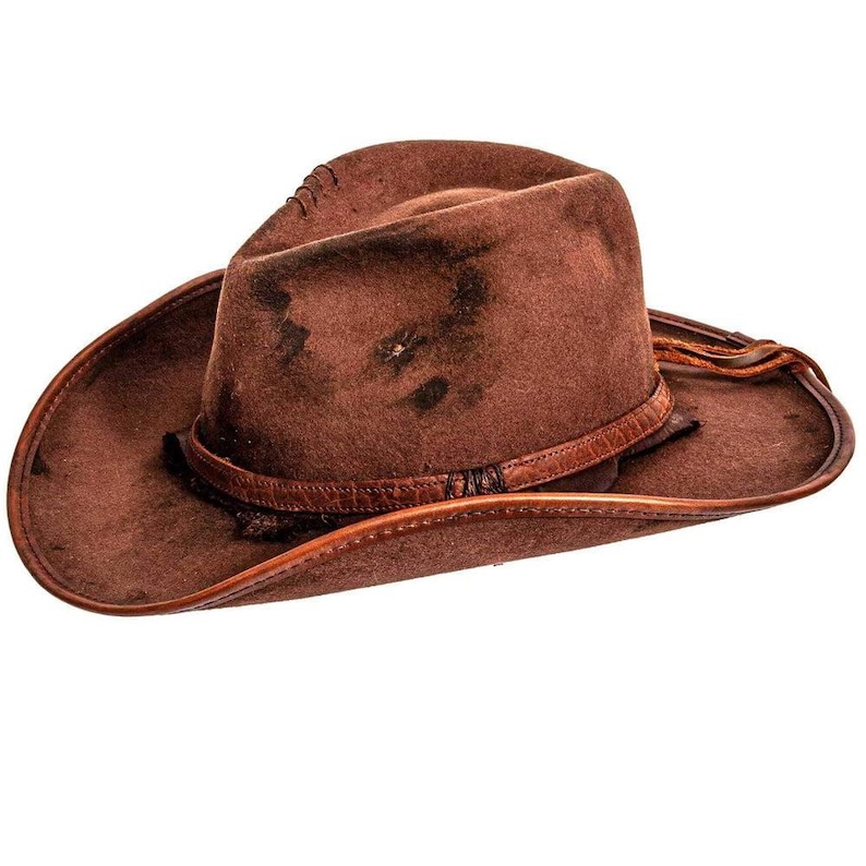 May include: A brown felt cowboy hat featuring a distressed finish with dark, weathered patches across the crown. The hat includes a thin, textured leather band encircling the base and a matching leather trim along the edge of the wide brim.
