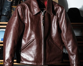 May include: A brown leather jacket with a zipper closure and a collared neckline hangs on a wooden hanger. The jacket has two front pockets and a red and black plaid lining. Other leather jackets and boots are visible in the background.