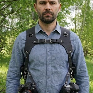 May include: A black leather dual camera harness featuring a textured finish, adjustable chest straps with metal buckles, and two secure attachment points for cameras. The harness is worn over a light blue button-down shirt, providing support for two cameras simultaneously.