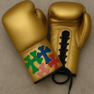 Puede incluir: Un par de guantes de boxeo dorados. Los guantes tienen cordones negros y diseños de cruces coloridas en verde, azul, rojo y rosa en la zona de la muñeca. Los guantes son de un material brillante.