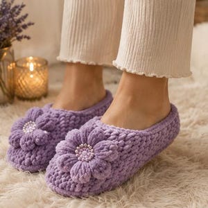 May include: A pair of lavender-colored slippers crafted from a soft, chunky knit yarn. Each slipper features a prominent, dimensional flower applique on the top, with a center embellished by small, light-colored beads for added texture.