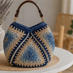 Op de afbeelding: Een ronde gehaakte handtas met een kleurverloop van donkerblauw, lichtblauw en beige garen. De tas heeft een donkerbruin gebogen handvat bevestigd met goudkleurige metalen kettingschakels en een V-vormige opening aan de bovenkant met textuur.