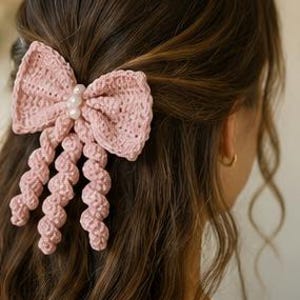 May include: A dusty pink crochet hair bow featuring a textured, knotted fabric construction with three dangling spiral-shaped ribbons. The centre of the bow is embellished with three small, round, white faux pearls arranged in a vertical line.