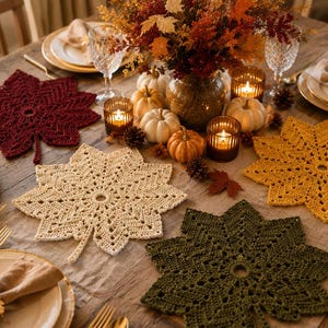 May include: A set of four crocheted leaf-shaped placemats in deep red, cream, mustard yellow, and olive green. Each placemat features an intricate, openwork stitch pattern radiating from a central ring, mimicking the veins and lobes of a maple leaf.