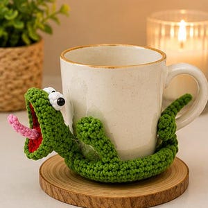 Puede incluir: Una taza de cerámica color crema envuelta en una funda de ganchillo con forma de lagarto verde. El lagarto tiene la boca muy abierta con un interior rojo, una lengua rosa que sobresale y dos ojos blancos con pupilas negras.