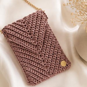 May include: A dusty rose crochet crossbody bag featuring a chevron pattern, a gold-tone metal chain strap, and a small circular gold-tone metal accent on the bottom right corner. The bag has a rectangular shape with a fold-over flap closure.