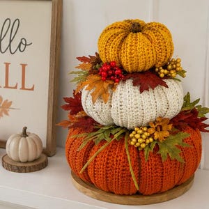Peut inclure: Un trio empilé de citrouilles au crochet en jaune, blanc et orange, décoré de feuilles d'automne artificielles, de baies rouges et d'accents dorés. Les citrouilles reposent sur une base en bois ronde, avec deux citrouilles plus petites à côté.