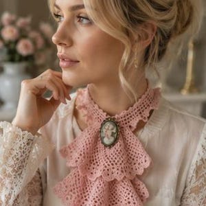 Puede incluir: Un volante de cuello tejido a ganchillo en color rosa empolvado con un diseño de niveles y bordes festoneados. El accesorio se sujeta en el centro con un broche ovalado que muestra una ilustración de retrato detallada en un marco bronce.