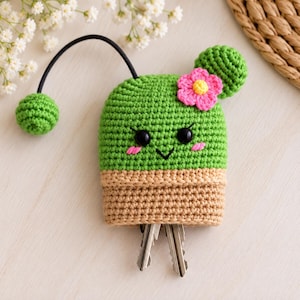 May include: A green crochet cactus key cover with a tan pot base, featuring a pink flower, black bead eyes, and pink stitched cheeks. A black elastic cord with a green crochet ball is attached to the top, and two metal keys hang from the bottom.