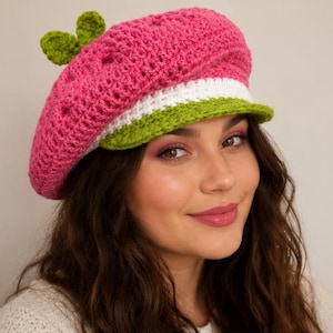 May include: A crocheted newsboy cap featuring a vibrant pink main body with a textured stitch pattern, a white band, and a green brim. Two small green crocheted leaves are attached to the top of the cap, adding a decorative detail to the design.